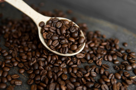 Fresh roasted arabica coffee beans in a wooden spoon and scattered coffee beans on a wooden table.