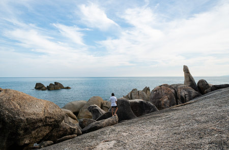 Young Man with a long hair in a white t-shirt, standing on a rock and enjoying a view on a sea....