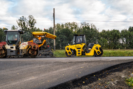 KYIV, UKRAINE - September 25, 2020:: Heavy asphalt road roller with heavy vibration roller compactor that press new hot asphalt and asphalt paver machine on a road construction site on a street.のeditorial素材