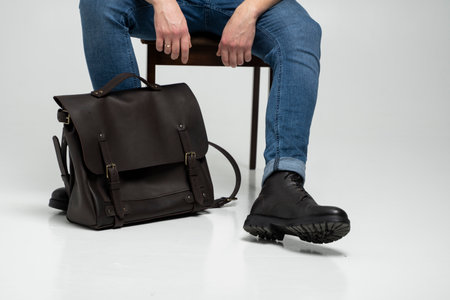 Man in a blue jeans and black boots sits on a chair with a brown mens shoulder leather bag for a documents and laptop on a white floor. Mens leather satchel, messenger bags, handmade briefcase.の写真素材
