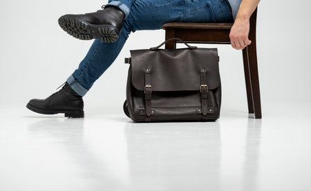 Man in a blue jeans and black boots sits on a chair with a brown mens shoulder leather bag for a documents and laptop on a white floor. Mens leather satchel, messenger bags, handmade briefcase.の写真素材