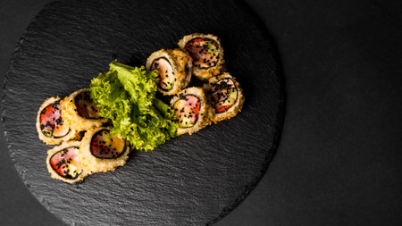 Custom sushi roll in tempura with nori, fresh salmon, tuna, avocado, masago caviar, drizzled with pineapple sauce with salad pouring as decoration on a black plate on a black table and background.の写真素材