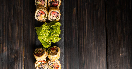 Custom sushi roll in tempura with nori, fresh salmon, tuna, avocado, masago caviar, drizzled with pineapple sauce with salad pouring as decoration on a black plate on a wooden table and background.の写真素材