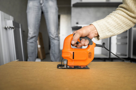 Man hand using electric jigsaw for cutting a fiberboard on a back side of a custom kitchen cabinet....