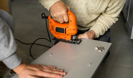 Man hand using electric jigsaw for cutting a fiberboard on a back side of a custom kitchen cabinet....
