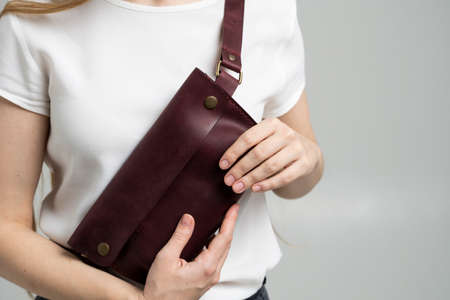 Girl in a white blouse with a leather red handmade bag over her shoulder. Designer dark red banana bag. Woman in a studio. Comfortable small bag for walking.の写真素材