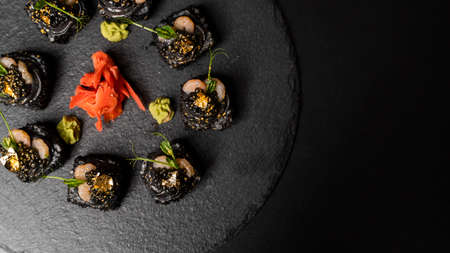 Sushi roll with nori, black rice, crab meat, cucumber, avocado, smoked salmon mousse, oar caviar, masago, shrimp cocktail and edible gold leaf with red ginger and wasabi on black table and background.の写真素材