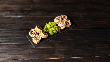 Custom sushi roll in tempura with nori, fresh salmon, tuna, avocado, masago caviar, drizzled with pineapple sauce with salad pouring as decoration on a black plate on a wooden table and background.の写真素材