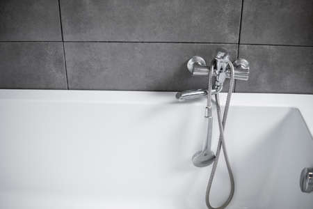 Chrome tap with a shower with a white bath in a bathroom with a grey tile. Stylish white bath with a silver tap. Minimalism in interior of bathroom.の写真素材