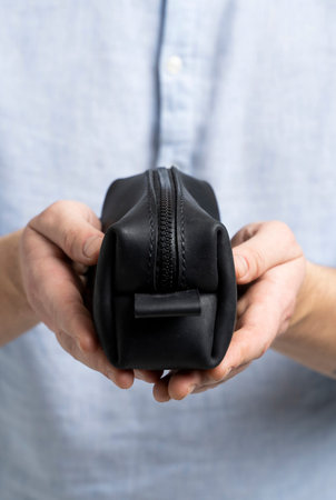 Mans black leather personal cosmetic bag or pouch for toiletry accessory in a mens hands in blue shirt. Style, retro, fashion, vintage and elegance.の写真素材