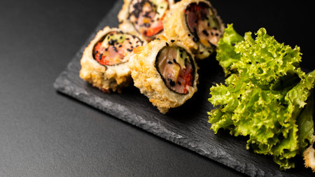 Custom sushi roll in tempura with nori, fresh salmon, tuna, avocado, masago caviar, drizzled with pineapple sauce with salad pouring as decoration on a black plate on a black table and background.の写真素材