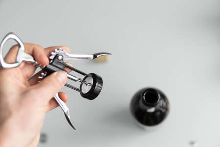 Metal corkscrew in a mans hand with a bottle of red wine on a background with a light white kitchen. Wine lover. Wine opener.の写真素材