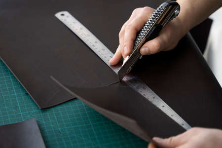Mens hand holding a stationery knife and metal ruler and cutting on a pieces for a leather wallet in his workshop. Working process with a brown natural leather. Craftsman holding a crafting tools.の写真素材