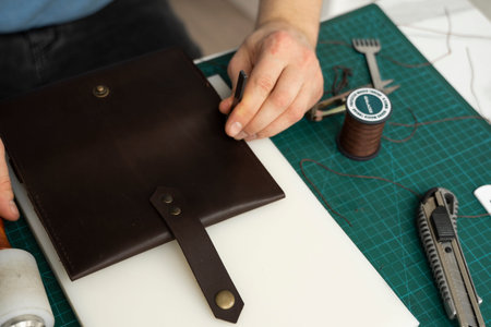 Mens hand working on the leather wallet in his workshop. Working process with a brown natural...