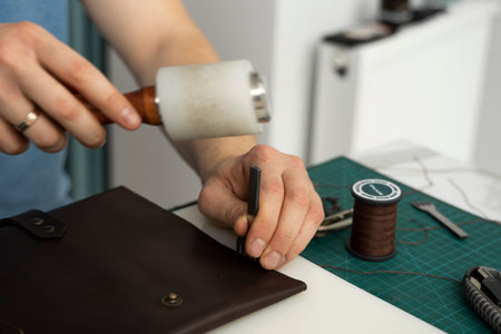 Mens hand holding a leather carving hammer and steel chisel hole punch and makes a leather wallet in...