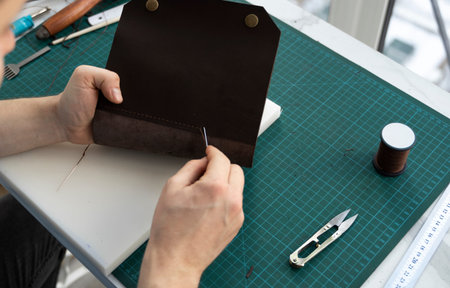 Mens hand working on the leather wallet in his workshop. Working process with a brown natural...