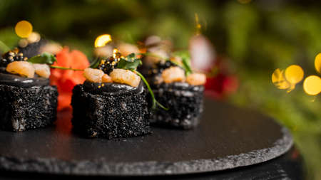Sushi with black rice, crab meat, avocado, smoked salmon mousse, oar caviar, masago, shrimp cocktail and edible gold leaf with ginger on black table for Christmas with a Christmas tree on background.の写真素材