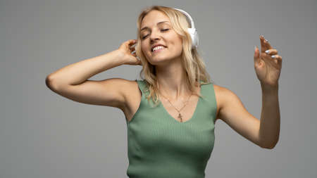 Beautiful attractive young blond woman wearing green t-shirt and glasses in white headphones listening music and smiling on grey background in studio. Relaxing and enjoying. Lifestyle.の写真素材