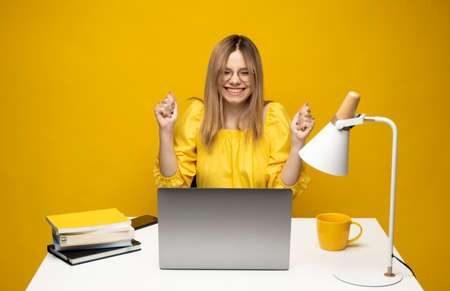 Excited young woman working with a grey laptop computer, notebook while sitting at the table with grey laptop. Smiling business woman or student received a good news isolated on a yellow background.の写真素材