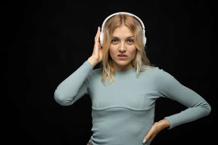Beautiful attractive young blond woman wearing blue t-shirt and glasses in white headphones listening music and smiling on black background in studio. Relaxing and enjoying. Lifestyle.の写真素材