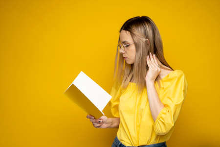 Beautiful smart young girl holding and reading book isolated on the yellow background. Portrait of attractive woman in a yellow blouse and wearing glasses reading book. Education, studying, knowledge.の写真素材