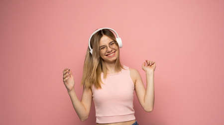 Beautiful attractive young blond woman wearing pink t-shirt and glasses in white headphones listening music and dancing on pink background in studio. Relaxing and enjoying. Lifestyle.の写真素材
