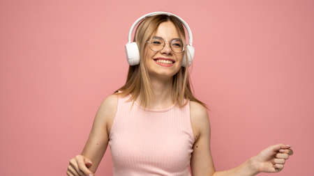 Pretty girl in a pink t-shirt and glasses listening music with her headphones and dancing on pink background.の写真素材