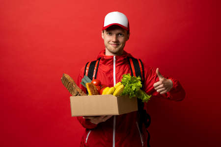 Happy courier man in cap, jacket and thermal bag backpack holds paper box with food products and showing a thumb up isolated on red background studio. Delivery service from groceries market to home.の写真素材
