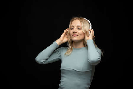Beautiful attractive young blond woman wearing blue t-shirt and glasses in white headphones listening music and smiling on black background in studio. Relaxing and enjoying. Lifestyle.の写真素材