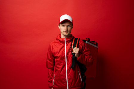 Young courier in a red uniform t-shirt and with red food thermo bag on a shoulder standing isolated...