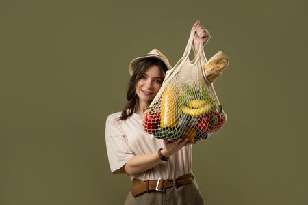 Portrait of smiling young woman in oversize t-shirt holding reusable string bag with groceries....