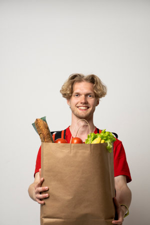 Portrait of pleased delivery man in red uniform smiling while carrying paper bag with food products isolated over white background.の写真素材