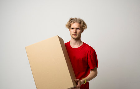 Male employee in in red uniform working as courier dealer and holds cardboard box isolated on white background, studio portrait. Service concept. Mock up copy space.の写真素材