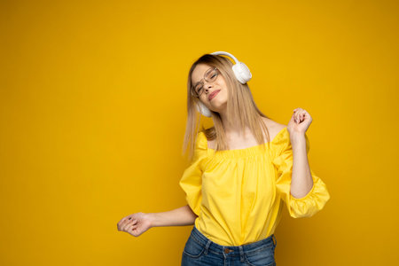 Happy girl in a glasses and yellow shirt dancing and listening to the music isolated on a yellow...