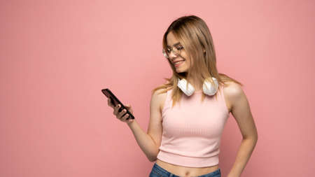 Surprised young happy girl in pink t-shirt using mobile phone, typing sms message isolated on pink background, studio portrait. People lifestyle concept. Mock up copy space.の写真素材