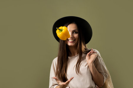 Eco friendly smiling woman in beige t-shirt and black hat throw up a yellow pepper in a air by one hand and holding reusable cotton eco bags full of groceries on a shoulder.の写真素材