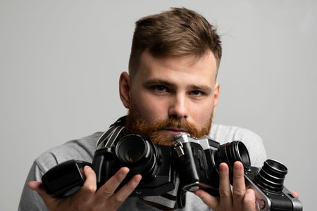 Bearded professional photographer in a grey t-shirt with a bunch of different cameras in a hands looking on a camera and ready for make a good shoot.の写真素材