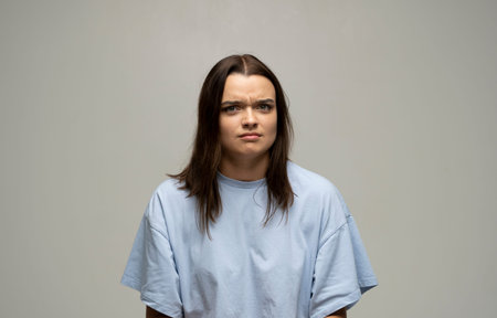 Portrait of an upset, angry young brunette girl in a blue oversize t-shirt looking in a camera...