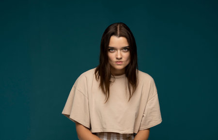 Portrait of an upset, angry young brunette girl in a beige oversize t-shirt looking in a camera...