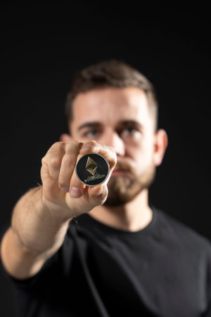Mature bearded man wearing in black t-shirt holding ethereum coin in hand and standing isolated on...