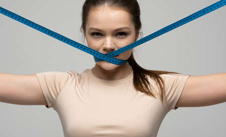 Young brunette woman in a beige t-shirt holding measuring tape around her mouth on white background. Healthy lifestyle concept.の写真素材