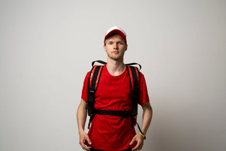 Young courier in a red uniform t-shirt and with red food thermo bag on a shoulder standing isolated...