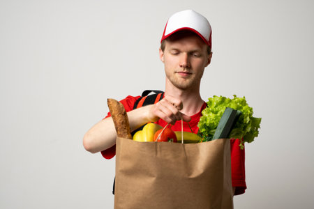 Grocery delivery courier man in a red cap and t-shirt with a thermal bag on a shoulders holds paper bag with food on white studio background. Food delivery service, online shopping.の写真素材