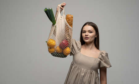 Brunette woman showing a reusable mesh shopping bag full of fresh groceries. Zero waste. Ecologically and environmentally friendly packets. Canvas and linen fabrics. Save nature concept.の写真素材