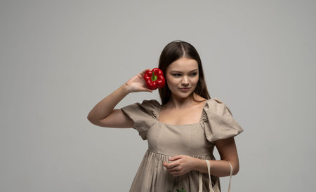Portrait of Charming brunette woman in a beige dress with cotton mesh eco bag and a red pepper holds near a face. Zero waste shopping concept.の写真素材