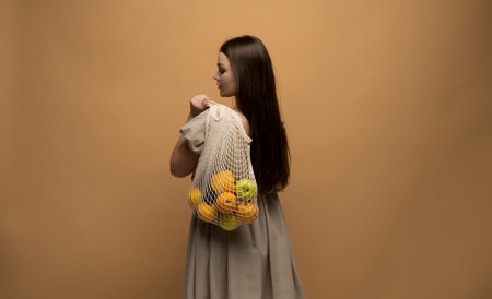 Back of woman in beige dress holding string shopping reusable mesh bag with fruits on a shoulder. Zero waste concept. Space for text. Reusable eco friendly object. Copy space.の写真素材