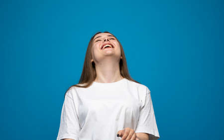 Young beautiful cute cheerful brunette girl in white t-shirt smiling looking at camera and showing a tongue over blue background studio. Emotions.の写真素材