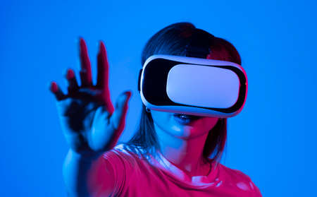 Excited brunette woman in white t-shirt getting experience using VR-headset glasses of virtual reality much gesticulating hands in neon light.の写真素材