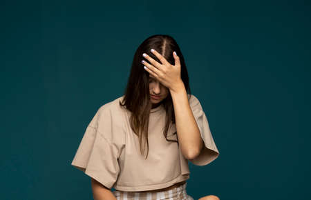 Portrait of stressed beautiful brunette woman in a beige t-shirt holding her head with hands having headache over cyan wall. Attractive female suffering from head pain.の写真素材