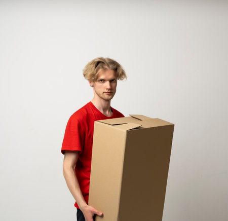 Portrait of a happy young delivery man in red uniform standing with parcel post box isolated over white background.の写真素材